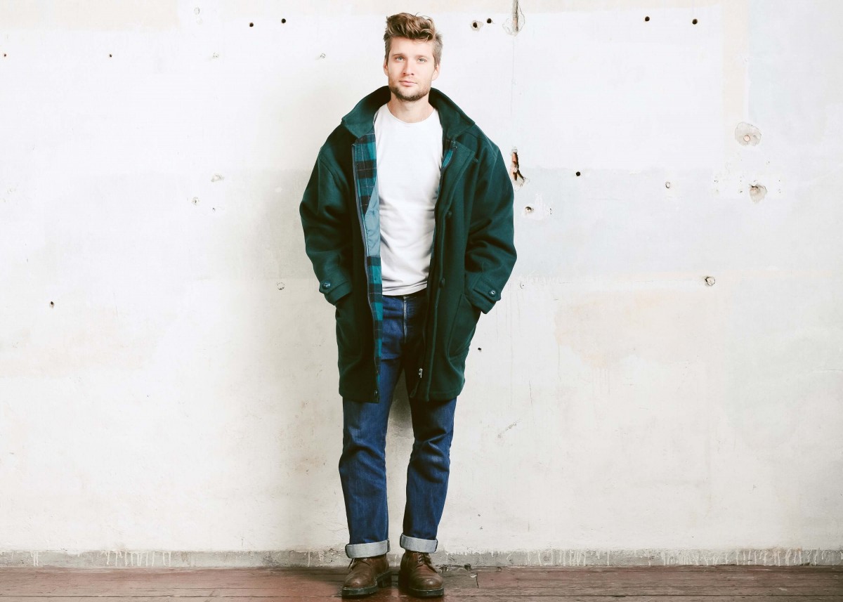 Pria mengenakan kaus putih polos dan jeans biru tua, dilapisi dengan overcoats berwarna hijau, sedang berdiri di depan dinding putih.